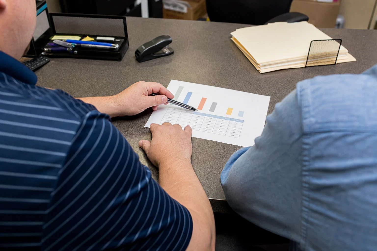 Agent in a striped polo points a pen at a printed bar chart and coverage table on an office desk while a client in a blue shirt looks on