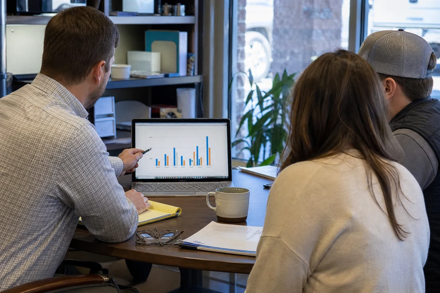 Agent in a checked button-down points to a bar chart on a laptop while a couple reviews it with him at a round table in a storefront office