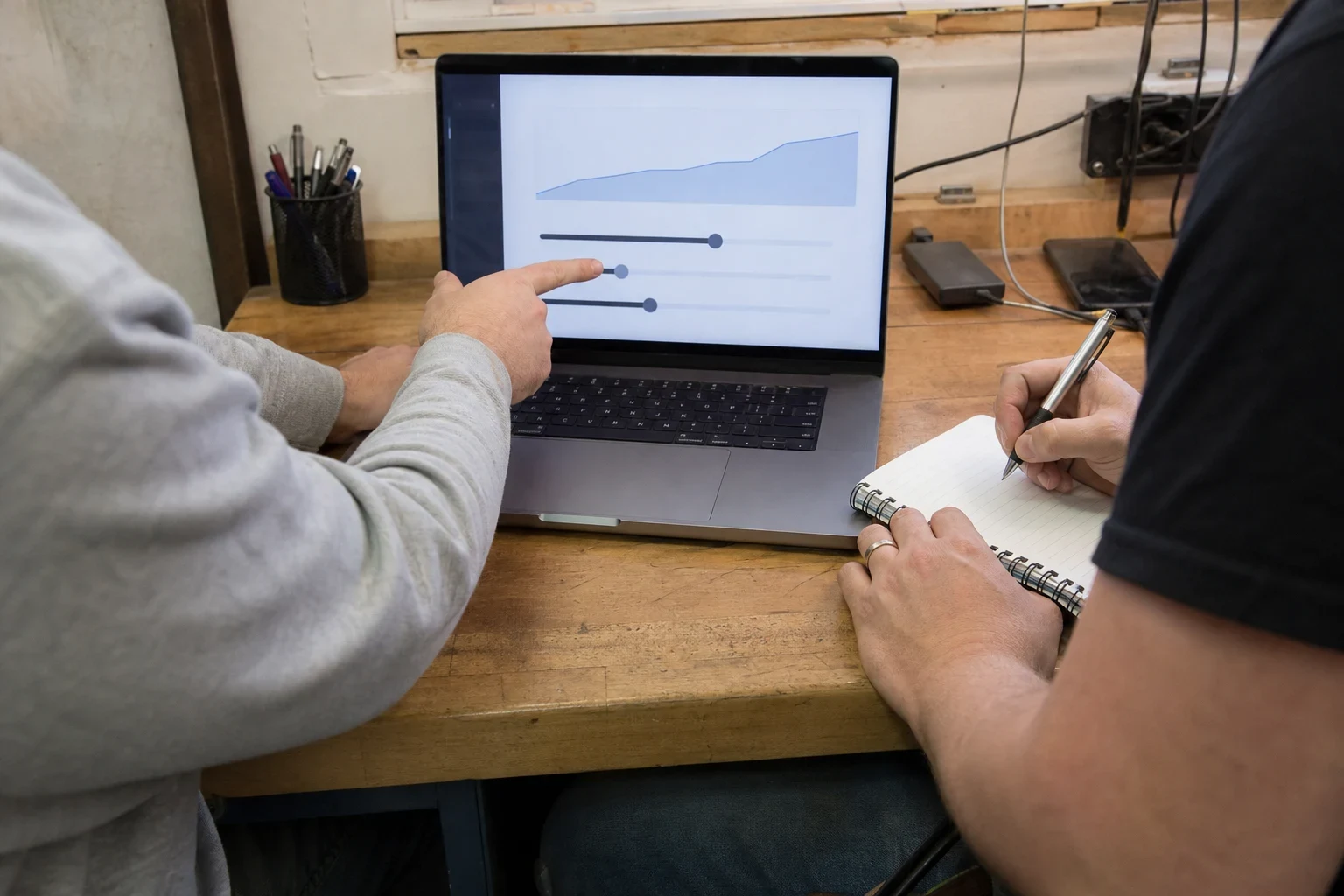 Person in a gray sweater points to sliders and a graph on a laptop while another person in a black T-shirt takes notes in a spiral notebook at a wooden desk