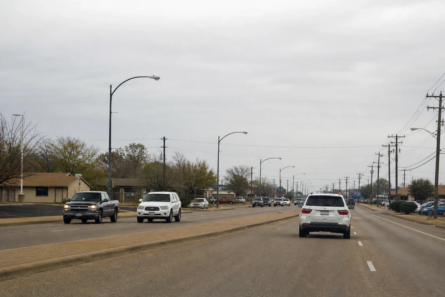 Wide divided street on an overcast day with pickups and SUVs in traffic, streetlights, utility poles, bare trees and low single-story buildings along the roadside