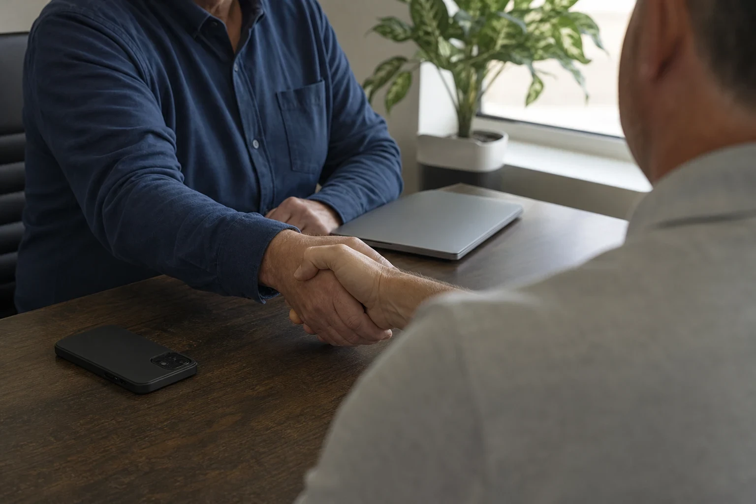Over-the-shoulder view of two men shaking hands across a wooden office desk with a phone, a closed laptop and a potted plant by a bright window